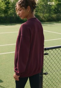 Burgundi värvi paelusiga dressipluus, millel on mugav istuvus ning ribatud mansetid ja ääred. Kantud mustade retuusidega, seistes rohelisel tennisekohal.