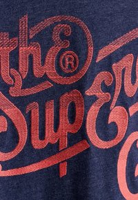 Tmavě modrá látka s texturovaným povrchem má velký červený kurzivní text, na kterém je napsáno "the Super." Design zahrnuje dekorativní ozdoby a symbol ochranné známky.
