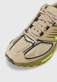 Μπεζ αθλητικά παπούτσια με διχτυωτό και συνθετικό επάνω τμήμα, που διαθέτουν μαύρες κορδόνες και πράσινες λεπτομέρειες. Ξεχωριστός σχεδιασμός σόλας με υφή.