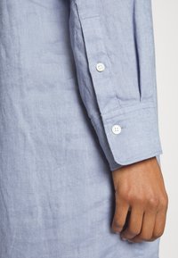 Prawe ręka zrelaksowana obok ciała, ubrana w jasnoniebieską koszulę z długim rękawem i białymi guzikami na mankiecie, na jednolitym tle.