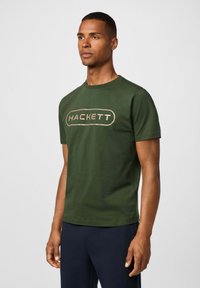 Camiseta de algodón verde con mangas cortas, que presenta un destacado logotipo beige de "HACKETT" en la parte frontal, cuello redondo y un diseño limpio.