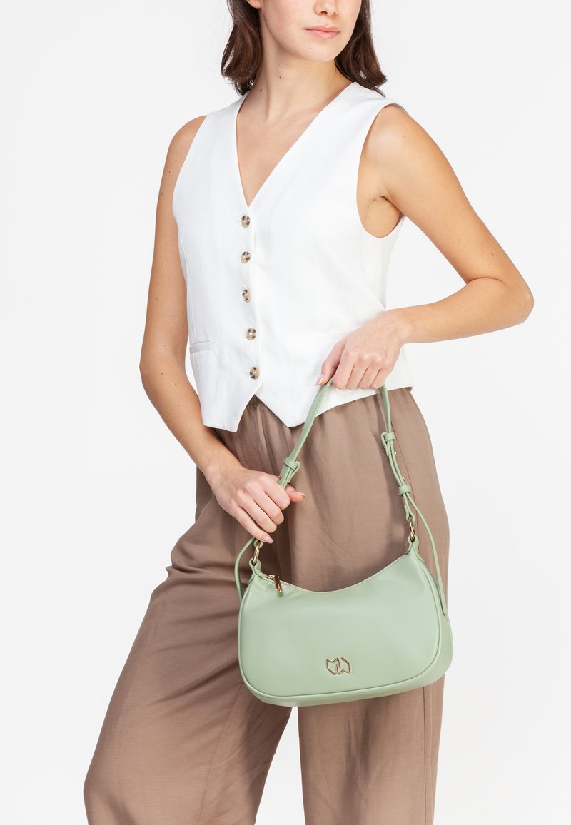 Grøn, buet håndtaske lavet af glat syntetisk materiale med en gylden logo accent, båret over et lysebrunt outfit med en tætsiddende hvid vest.
