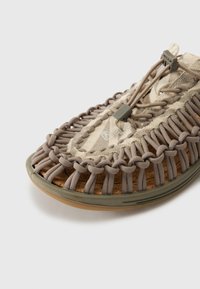 Sandal tertekstur yang menampilkan bagian atas tenun berwarna beige, tali berwarna abu-abu, insole gabus, dan outsole karet. Detailnya termasuk desain melingkar di sekitar jari kaki.