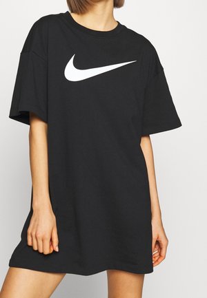 Černé bavlněné volné tričko s krátkými rukávy, na prsníku je velké bílé logo Nike ve tvaru šipky, minimalistický design.