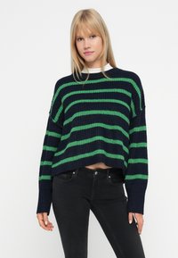 Mujer rubia vistiendo un suéter de punto rayado en azul marino y verde con jeans negros de cintura alta, de pie frente a un fondo blanco.