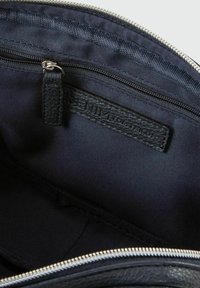 Interno di una borsa in pelle nera con una texture liscia, dotata di una tasca con zip e un'etichetta in denim. Il tessuto blu navy riveste l'interno.