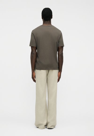 Άνδρας γυρισμένος από την πλάτη, φοράει καφέ κοντομάνικη μπλούζα και μπεζ παντελόνι με φαρδιές πλούσιες γραμμές, stand μπροστά σε απλό λευκό φόντο.