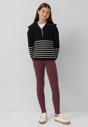 Schwarz-weißer gestreifter Strickpullover mit hohem Kragen und halbem Reißverschluss, kombiniert mit burgunderfarbenen Leggings und weißen Sneakers mit braunen Akzenten.