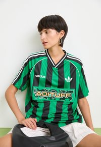 Žalias ir juodas dryžuotas trumpomis rankovėmis džersis su V formos iškirpte, kuriame yra didelis "ADITUFF" grafikas ir Adidas logotipas. Lygi audinio tekstūra.