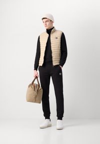 Beige steppelt mellény fekete garbó felett, fekete jogger nadrággal és fehér sportcipővel párosítva. Barna részletekkel rendelkező bézs táskát tart a kezében.