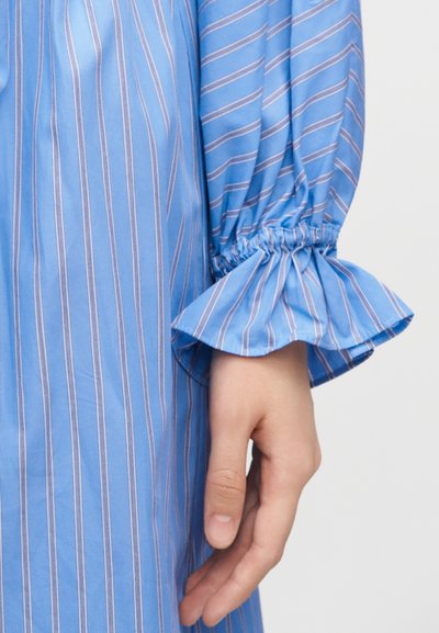 Χέρι με ανοιχτόχρωμο τόνο δέρματος δίπλα σε μπλε ύφασμα με λευκές κάθετες ρίγες και συλλεγμένο, φουσκωτό μανσέτα σε μακρύ μανίκι.