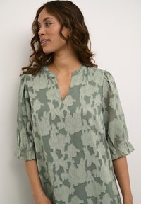 Ljusgrön blus med blommönster, med puffärmar och v-ringning. Tillverkad av ett texturerat, semi-genomskinligt tyg.
