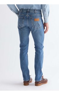 Férfi világoskék farmernadrágot és barna hajócipőt visel, háttal áll, kezei lazán az oldalán.