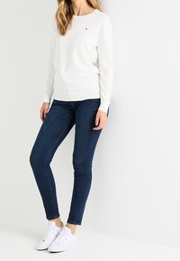 Valkoinen neulepaita pyöreällä kaula-aukolla, yhdistetty tummansinisiin kapeisiin farkkuihin ja valkoisiin tennareihin. Neuleessa hillitty logoyksityiskohta.