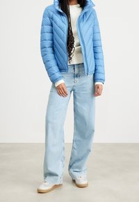 Světlomodrá péřová bunda s horizontálním prošitím, zapínáním na zip a kapucí, doplněná světle modrými širokými džínami a bílými teniskami.