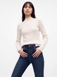 Ung kvinde iført en cremefarvet ribstrikket sweater og mørkeblå jeans, stående med hænderne nær lommerne mod en ensfarvet hvid baggrund.