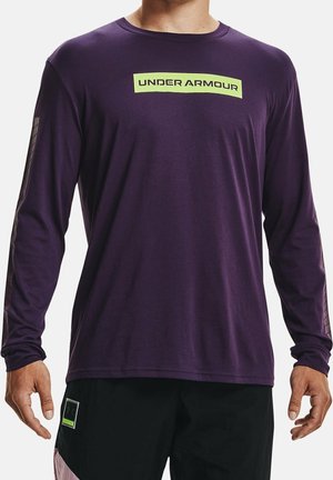 Mann trägt ein violettes Langarmshirt mit einem neongrünen Under Armour-Logo und schwarze Shorts mit einem kleinen Etikett am linken Bein.