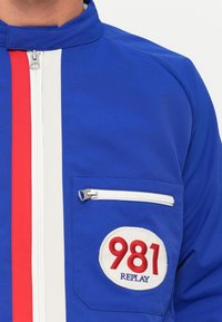 Blå jakke med lodrette blå og hvide striber, lynlås på brystlommen og en hvid lap med teksten "981 REPLAY" i røde og blå bogstaver.