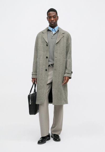 Sivi kaput s uzorkom na zube, zakopčan, nošen preko sivog džempera i plave košulje, u kombinaciji s bež hlačama i crnim cipelama, drži crnu torbu.