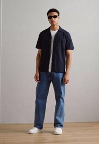 Camisa negra de manga corta con textura sobre una camiseta blanca, combinada con unos jeans azules holgados y zapatillas blancas. El modelo lleva gafas de sol oscuras.