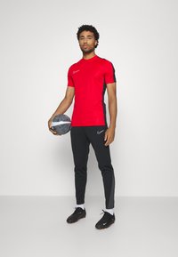 T-shirt de sport rouge avec des accents noirs, associé à un pantalon noir. Marque Nike sur le t-shirt et les chaussures. Modèle tenant un ballon de soccer.
