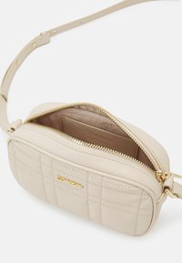 Béžová kabelka přes rameno z prošívané umělé kůže se zlatým zipem a logem. Obsahuje jeden prostor s hladkým vnitřkem.