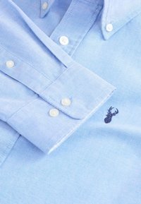 Hellblaues Hemd mit weißen Knöpfen und einem kleinen, dunkel gestickten Hirsch-Logo auf der Brust in der Nähe der Manschette.