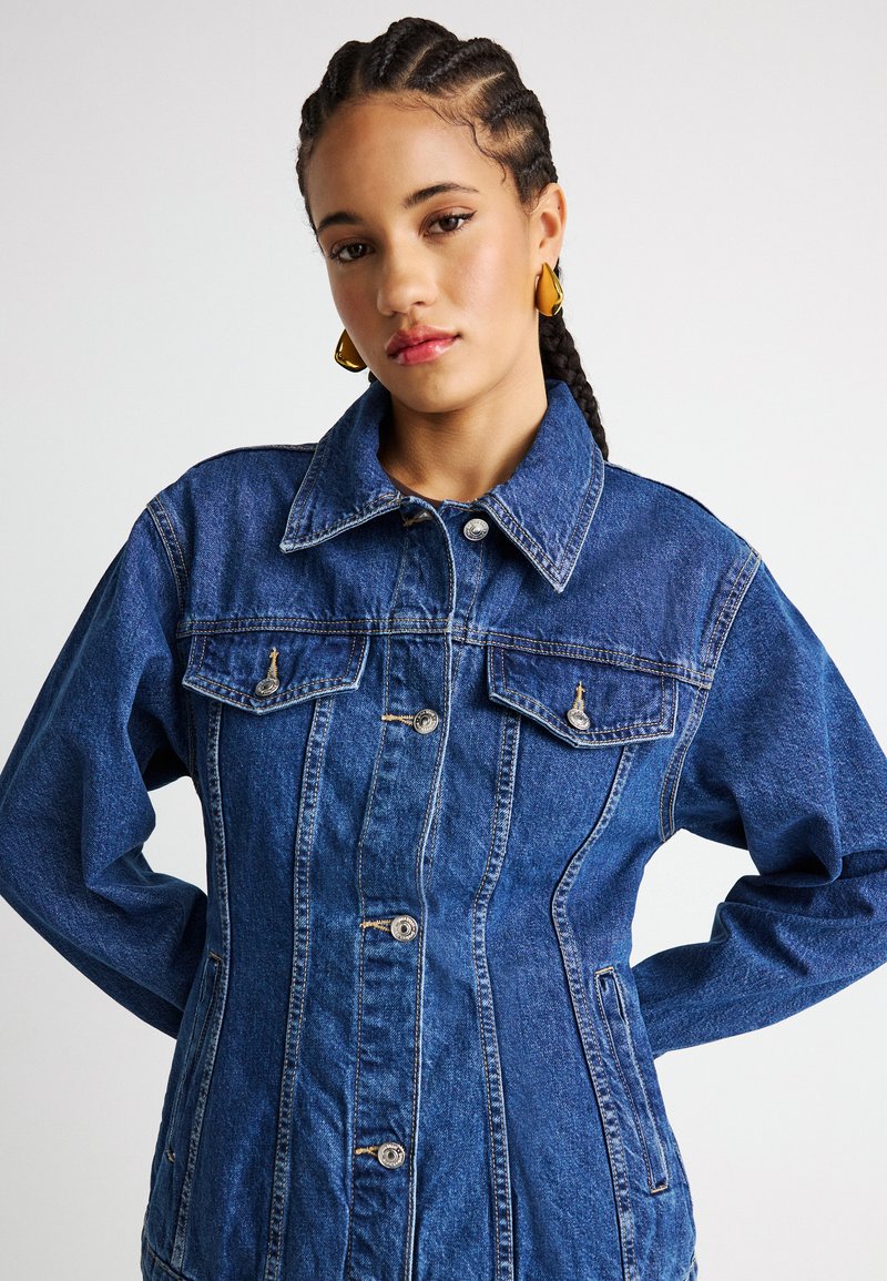 Νεαρή γυναίκα με πλεξούδες, φορώντας στενή μπλε τζιν μπουφάν με τσεπάκια στο στήθος με κουμπιά και χρυσά σκουλαρίκια, σε απλό φόντο.