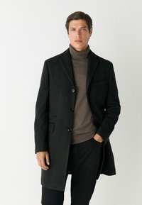 Mørk uldfrakke med notched lapels, tre knapper og en enkelt brystlomme, lagt over en brun striktrøje med høj hals.