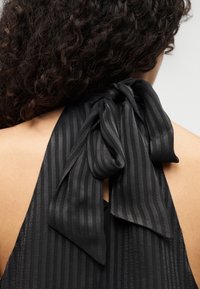 Μαύρο τοπ με halter neck και μεγάλο φιόγκο στο πίσω μέρος, με κάθετη υφή ριγέ και επίπεδη, λεία υφή υφάσματος.