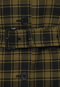 Mustast ja kollasest ruudulisest kangast, millel on suur nupp ja must vöö ristkülikukujulise pandlaga. Tekstuur on sile ja kudumismustriga.