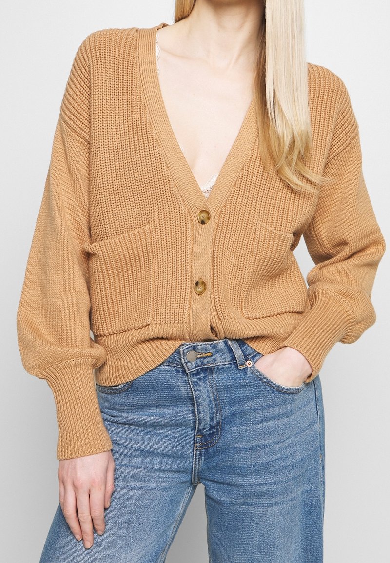 Knitted tan cardigan with a deep V-neck, long sleeves, two front pockets, button closure, and ribbed cuffs and hem. Paired with blue jeans.