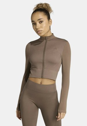 Giacca atletica corta marrone con zip, maniche lunghe e trama a coste, abbinata a leggings coordinati a vita alta. Dettaglio logo discreto.