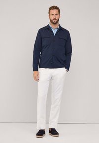 Granatowa koszula zapinana na guziki z dwiema kieszeniami na piersi, białe spodnie i czarne buty z plecioną podeszwą. Gładki materiał i dopasowany krój.