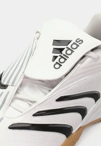 Sapatilha Adidas branca com um upper texturizado, acentos em preto, design riscado e sola de borracha. Logotipo proeminente na lingueta.