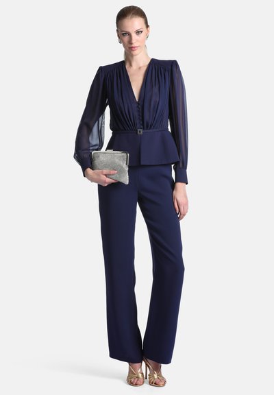 Femme portant un chemisier bleu marine à manches longues et col en V avec un pantalon assorti, tenant une pochette argentée et portant des talons ouverts dorés.