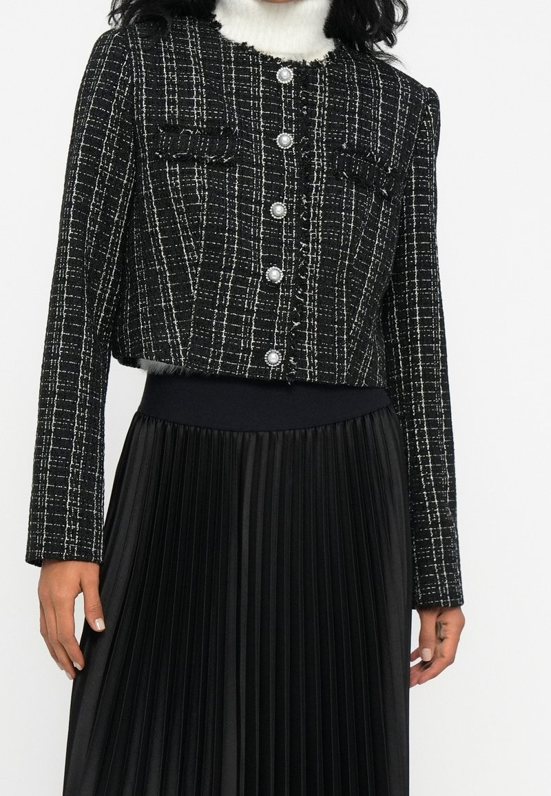 Femme portant une veste courte à carreaux noir et blanc avec des boutons en perle et une jupe plissée noire.