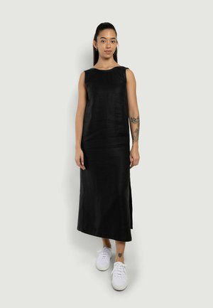 Donna in piedi che indossa un vestito midi nero senza maniche con spacchi laterali e sneakers bianche, mostrando tatuaggi floreali sul braccio destro e sulla caviglia sinistra.