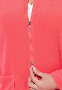 Chaqueta de cremallera de color coral con una textura suave. Presenta una cremallera metálica, bolsillos laterales y un corte relajado. Primer plano de la cremallera y la mano.