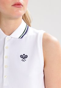 Bílý bezrukávový polo tričko s límečkem, s modro-zeleným proužkovaným akcentem a vyšívaným logem tenisové rakety. Viditelné knoflíky.