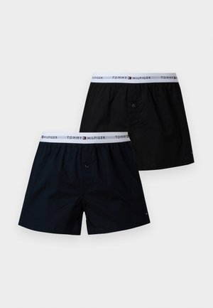 Høj, skjorteløs mand iført mørke shorts står barfodet ved siden af en mørkegrøn læderlænestol mod en almindelig lys væg.