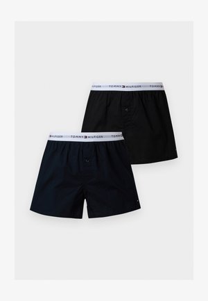 Ψηλός άνδρας χωρίς πουκάμισο με σκούρα σορτς στέκεται ξυπόλητος δίπλα σε σκούρο πράσινο δερμάτινο πολυθρόνα μπροστά σε έναν απλό φωτεινό τοίχο.