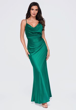 Femme aux longs cheveux foncés portant une robe en satin vert émeraude sans manches et des talons ouverts, debout devant un fond clair uni.