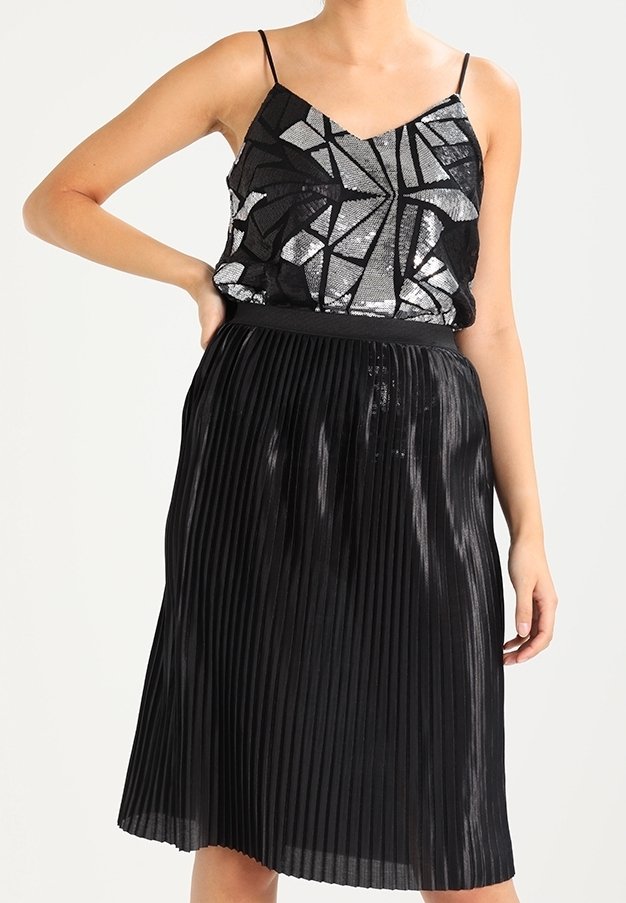 Jupe plissée noire à texture brillante associée à un débardeur noir et argenté à sequins avec un motif géométrique et des bretelles fines.