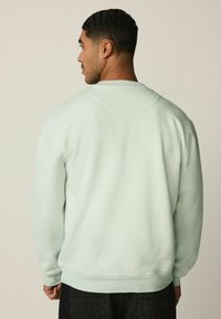 Hombre con cabello corto y rizado de espaldas, vestido con una sudadera verde clara y pantalones negros, frente a un fondo beige liso.