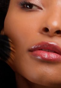 Gros plan d'une personne à la peau lisse appliquant du maquillage avec un pinceau sur la joue, lèvres brillantes avec un rouge à lèvres rouge, œil partiellement visible.