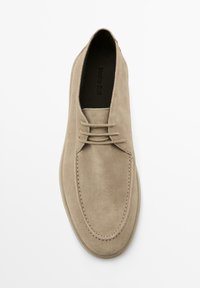 Chaussure en daim beige avec un bout rond, des lacets fins et des coutures le long des bords. Texture lisse et design minimaliste.