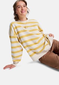 Gelb-weiß gestreifter Sweatshirt mit "Roxy Established 1990" in Weiß bedruckt; hat einen lässigen Schnitt und gerippte Bündchen, kombiniert mit braunen Cordhosen.