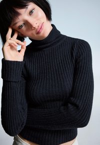Črna rebrasta jopica s turtleneck iz teksturirane tkanine, z oprijetim krojem in dolgimi rokavi, ki se končajo tik nad pasom.
