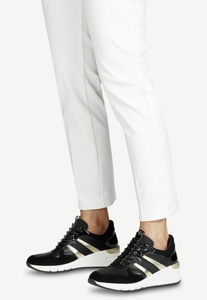 Des baskets noires et dorées présentent un dessus en maille, des panneaux texturés et une semelle blanche. Portées avec un pantalon blanc ajusté, mettant en valeur un design épuré.